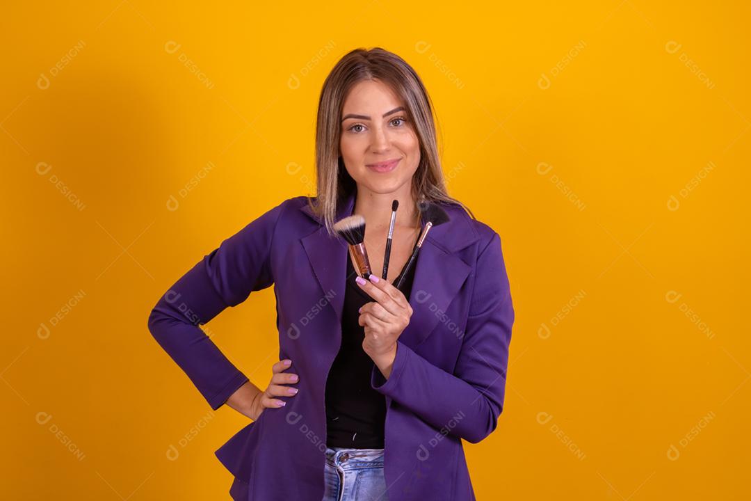 Mulher loira com um blazer roxo segurando pinceis de maquiagem