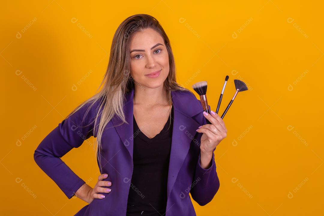 Mulher loira com um blazer roxo segurando pinceis de maquiagem
