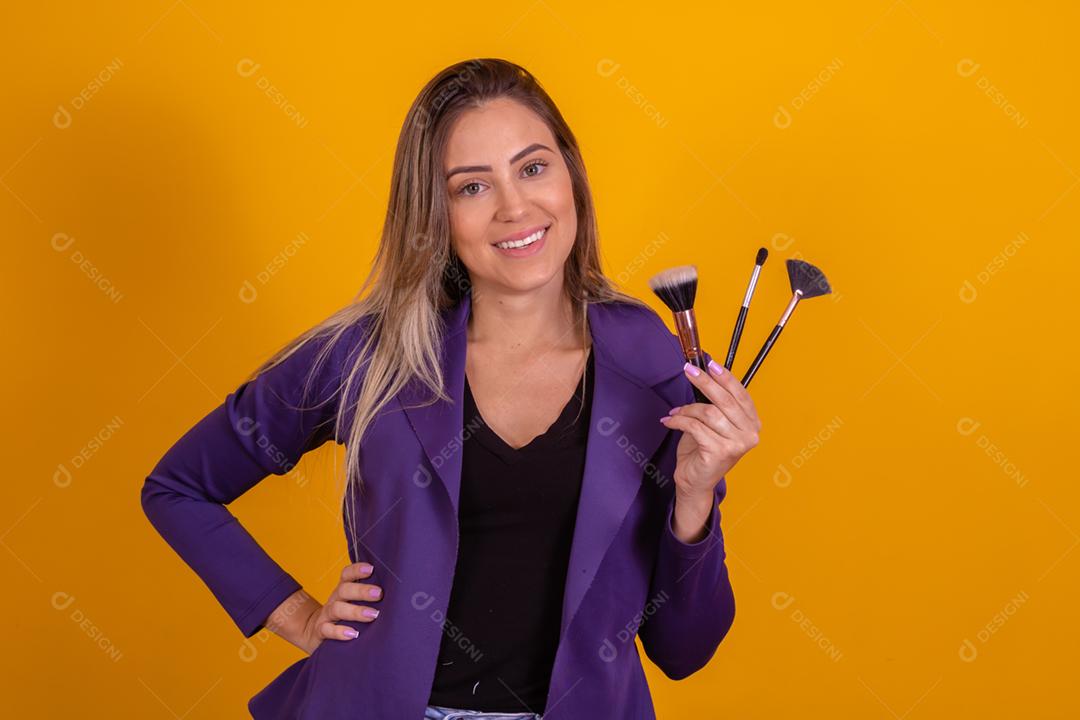 Mulher loira com um blazer roxo segurando pinceis de maquiagem