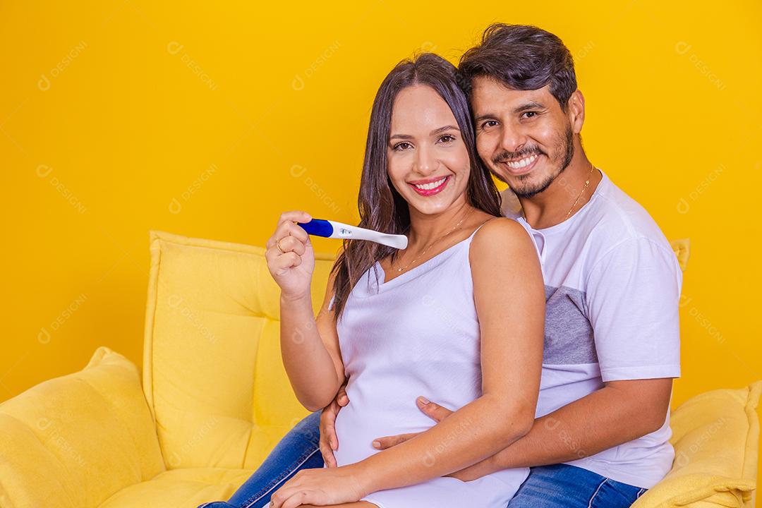 Feliz casal multinacional comemorando o resultado positivo do teste de gravidez no sofá em casa. Jovem abraçando