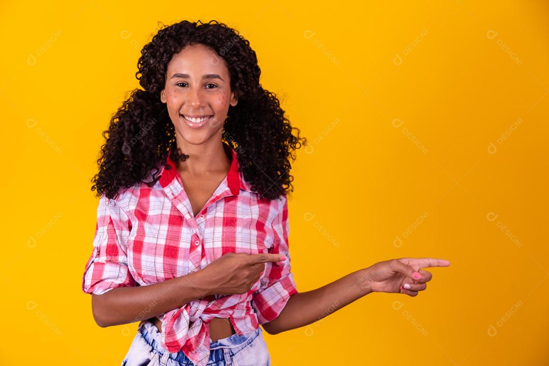 Mulher afro vestida com roupa de festa junina apontando para o lado.