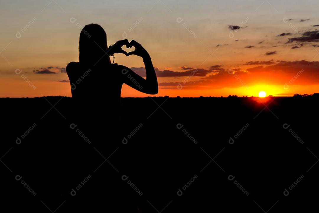 Belo pôr do sol com silhueta de menina fazendo coração com as mãos.