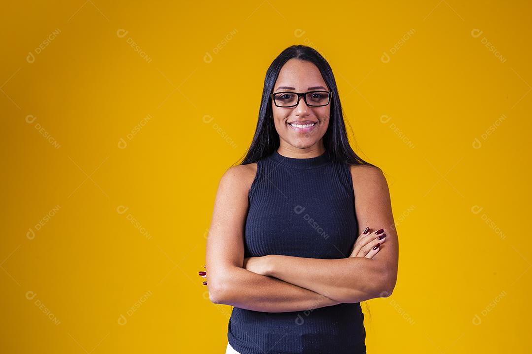 Mulher jovem e bonita com cabelo comprido, usando óculos e braços cruzados sobre fundo amarelo