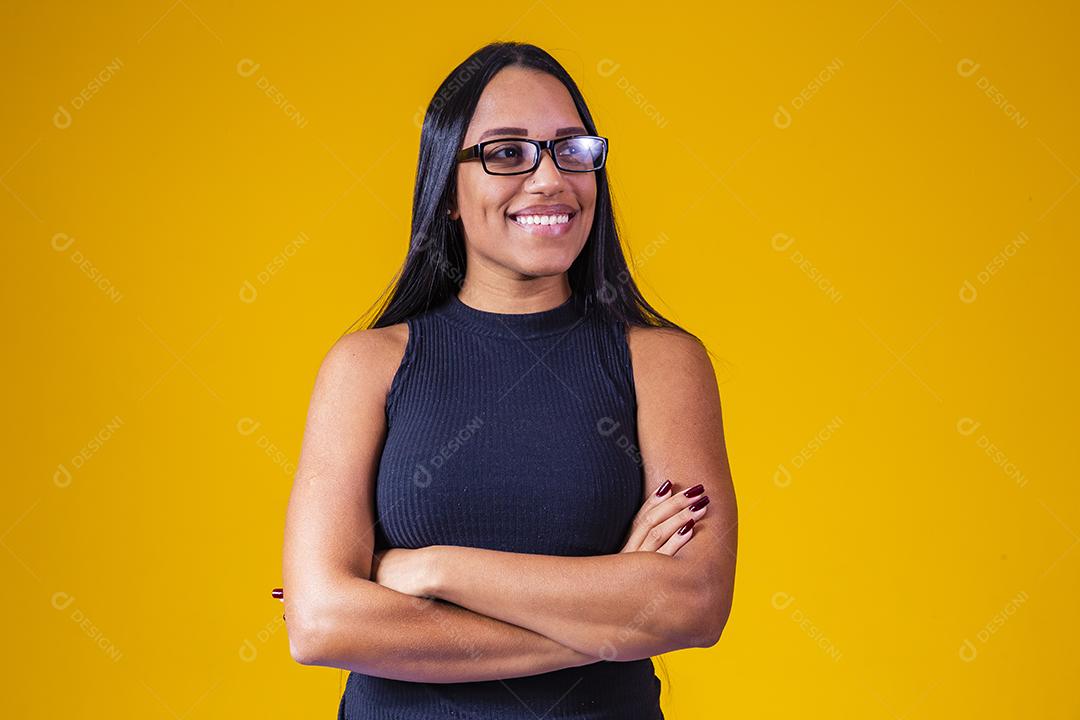 Mulher jovem e bonita com cabelo comprido, usando óculos e braços cruzados sobre fundo amarelo