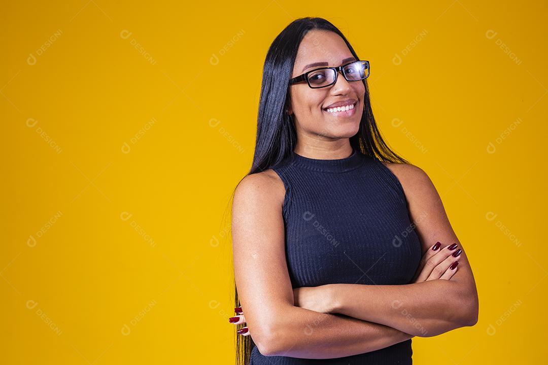 Mulher jovem e bonita com cabelo comprido, usando óculos e braços cruzados sobre fundo amarelo