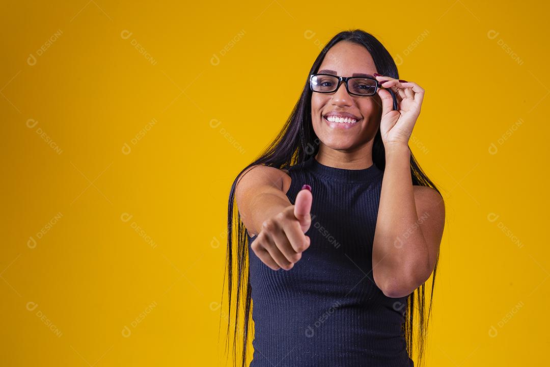 Mulher jovem e bonita com cabelo comprido, usando óculos e braços cruzados sobre fundo amarelo