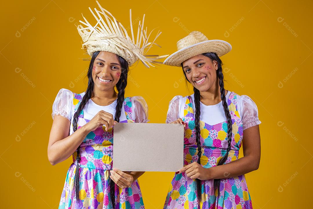 jovens irmãs gêmeas vestindo roupas tradicionais para Festa Junina segurando uma placa