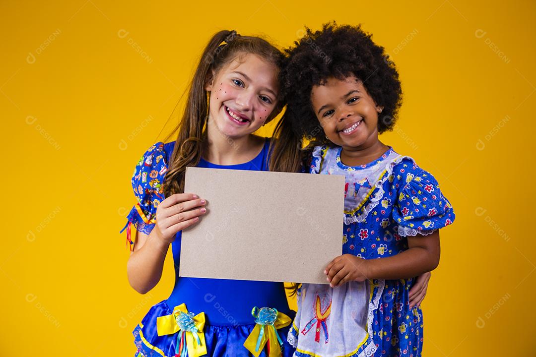 Duas crianças amigas com roupa de festa junina segurando um cartão em branco