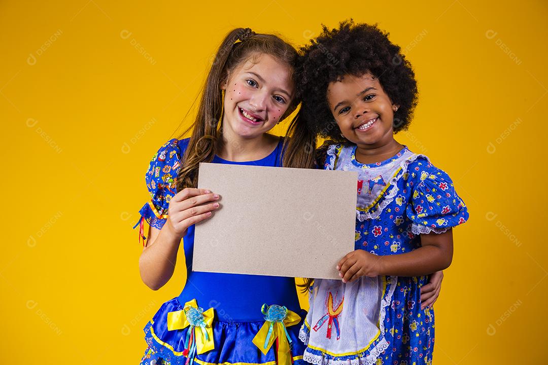 Duas crianças amigas com roupa de festa junina segurando um cartão em branco