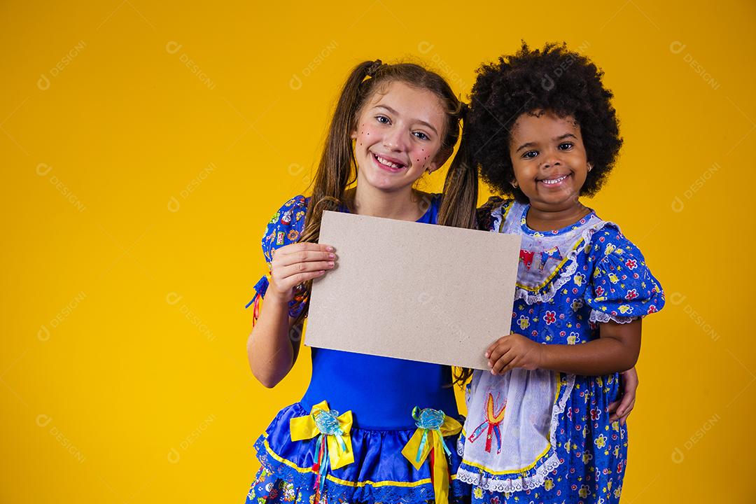 Duas crianças amigas com roupa de festa junina segurando um cartão em branco