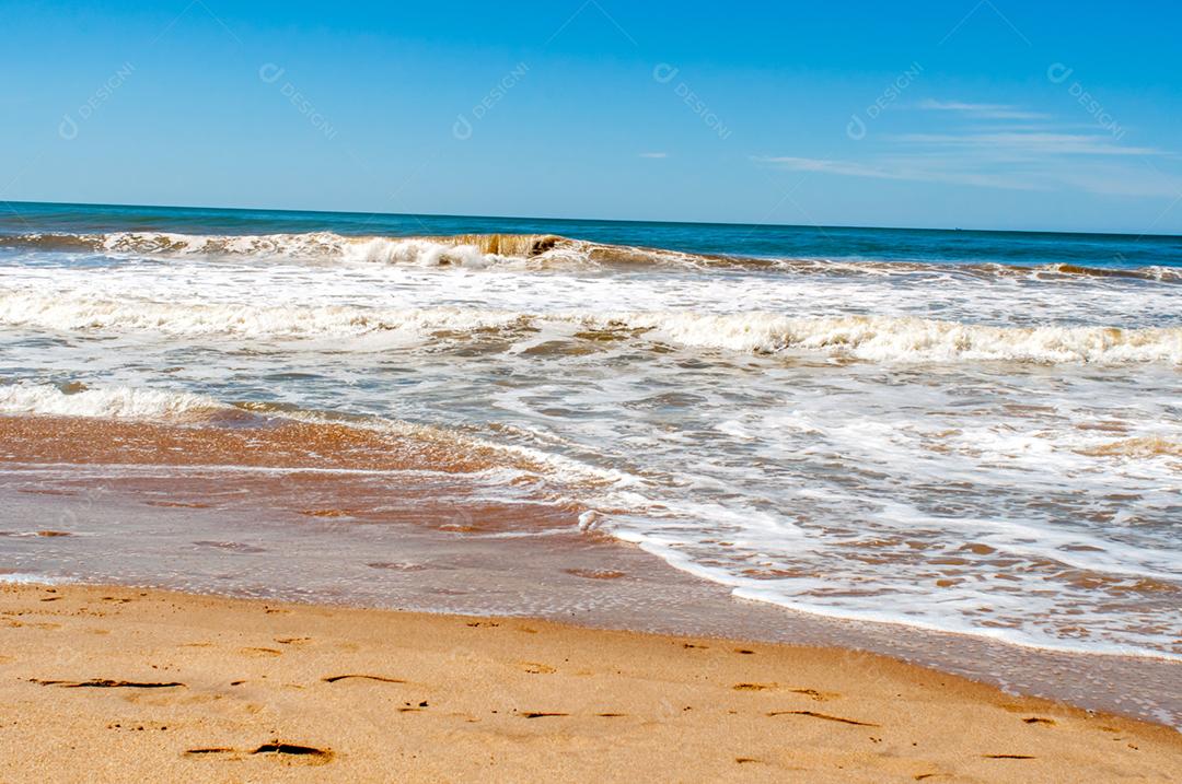 Praia em um dia ensolarado com céu azul, ondas caindo.