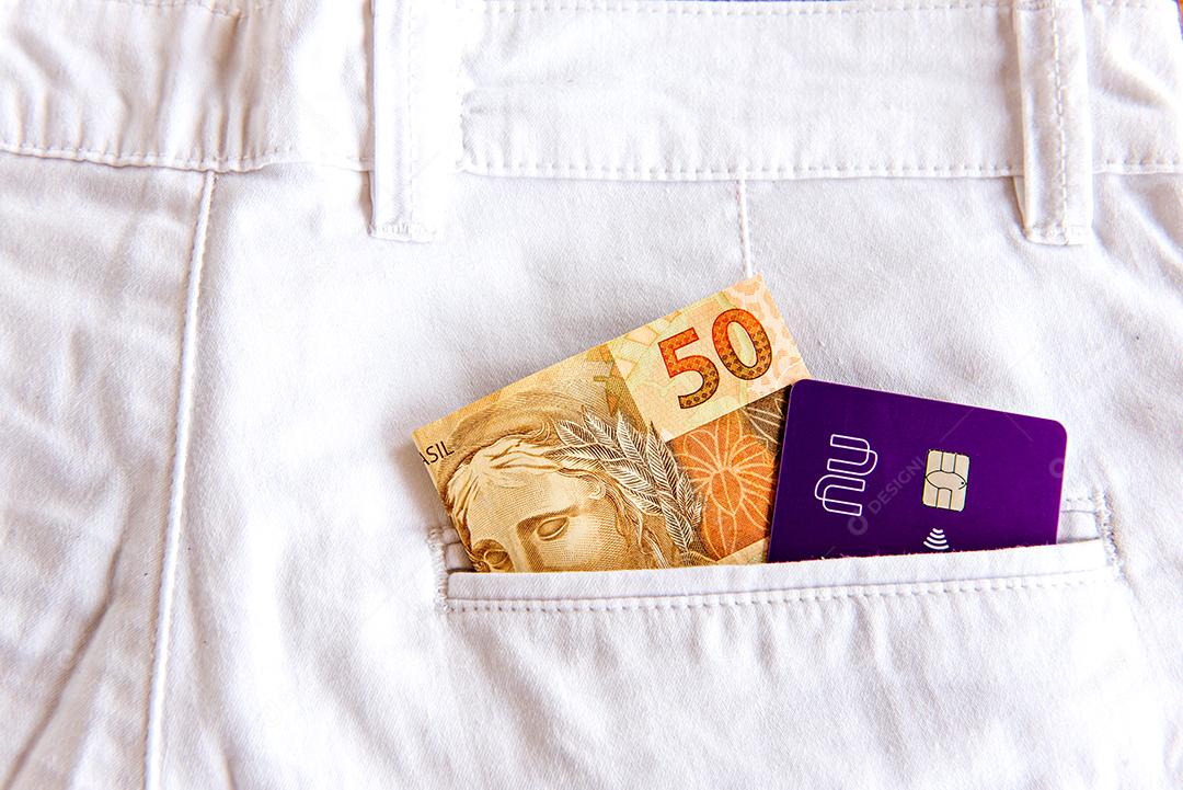 Dinheiro brasileiro e cartão de crédito Nubank no bolso da calça jeans branca.