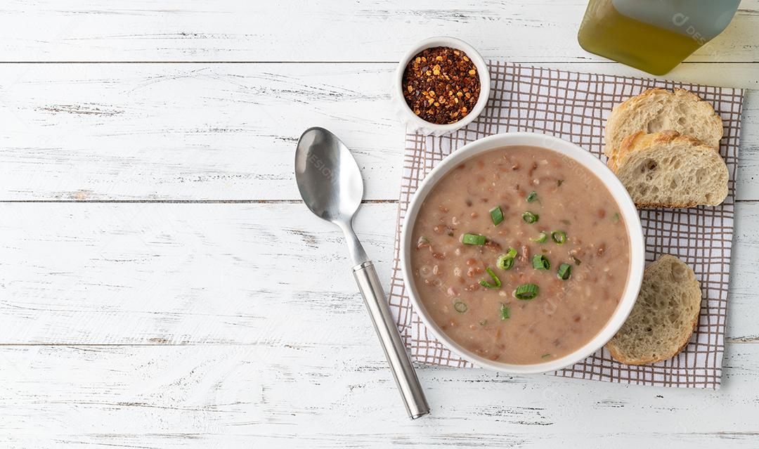 Sopa de feijão em uma tigela com fatias de pão e temperos sobre madeira