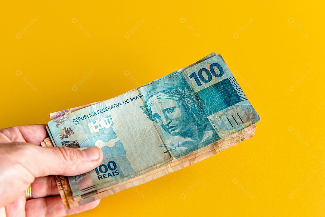 Mão segurando notas de dinheiro real brasileiro em fundo amarelo