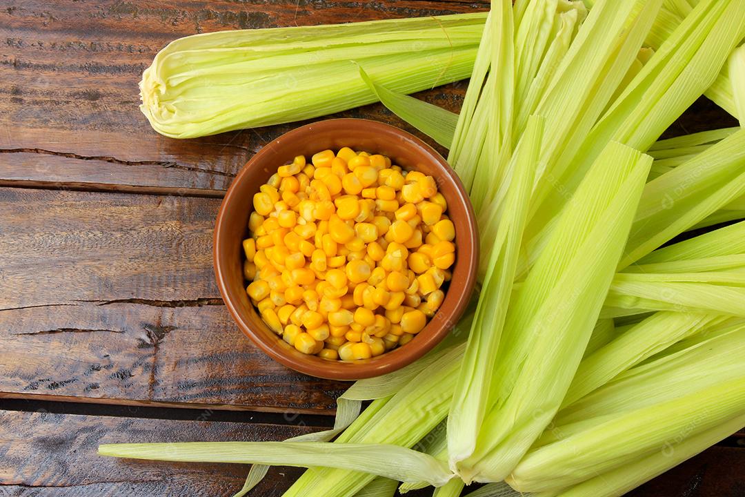 Grãos de milho cru, dentro da tigela de cerâmica, ao lado da espiga de milho na mesa de madeira rústica