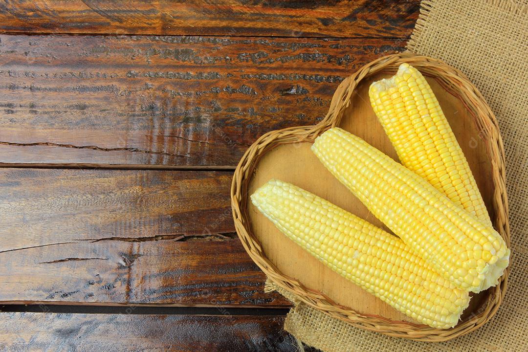 Espiga de milho, fresca, colhida da plantação dentro da cesta com forma de coração na mesa de madeira rústica