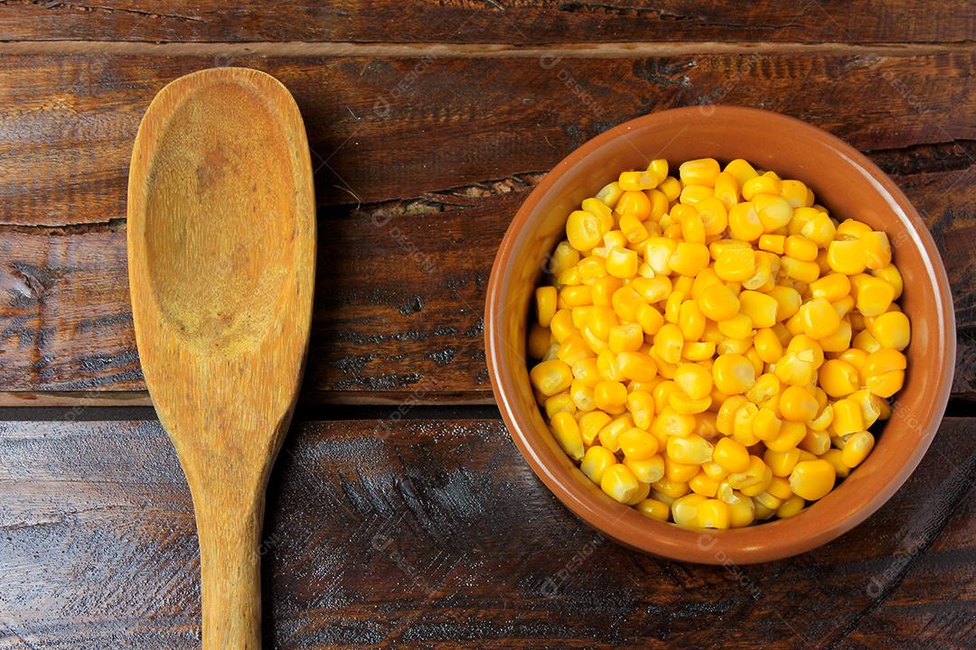 Grãos de milho cru, dentro da tigela de cerâmica, ao lado da colher de pau na mesa de madeira rústica