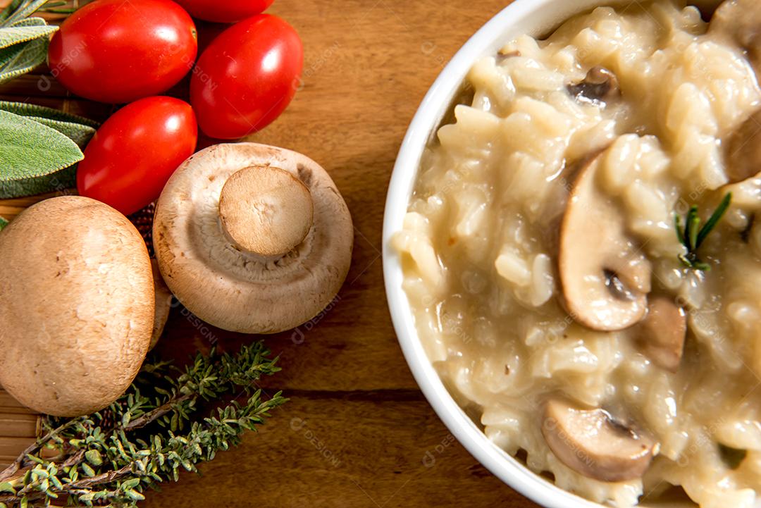 Tigela com delicioso risoto com cogumelo na mesa de madeira, tomate cereja, ervas e cogumelos