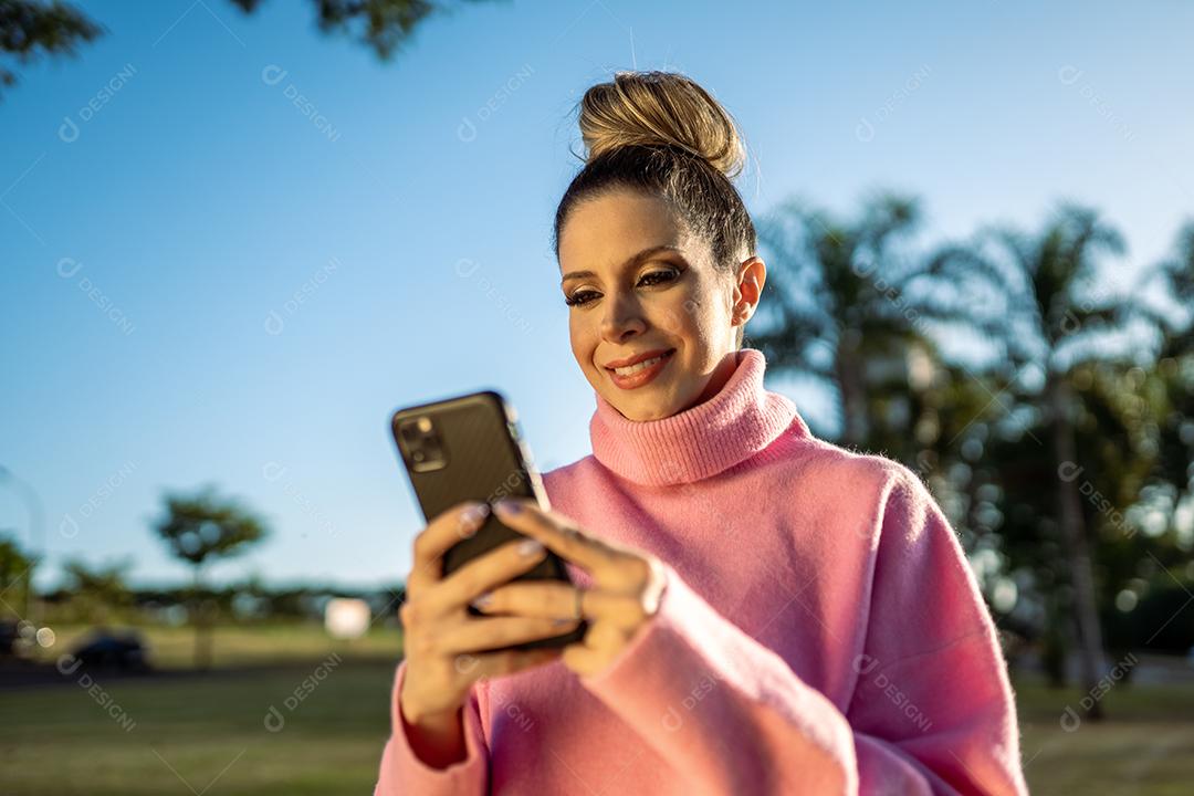Imagem de uma jovem loira latina animada feliz usando o celular no fundo do parque