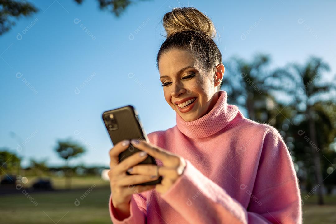 Imagem de uma jovem loira latina animada feliz usando o celular no fundo do parque