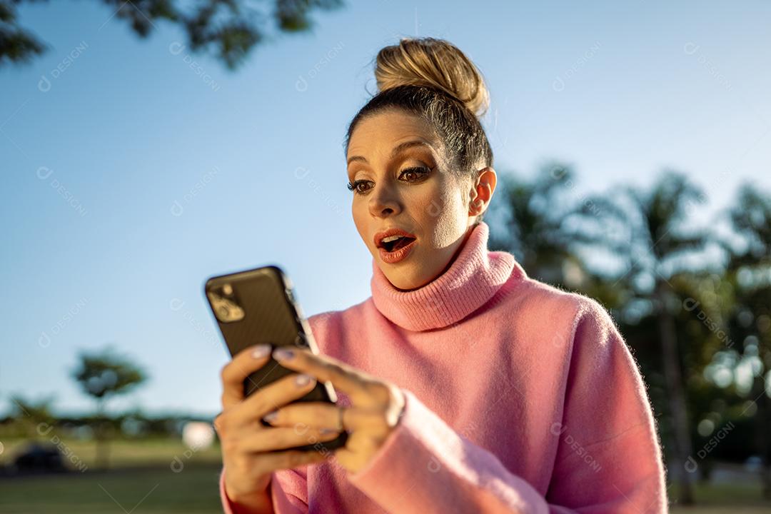 Imagem de uma jovem loira latina animada feliz usando o celular no fundo do parque