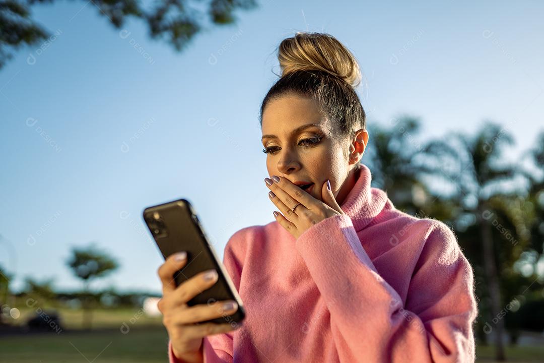 Imagem de uma jovem loira latina animada fazendo gesto de vencedor usando o celular no fundo do parque