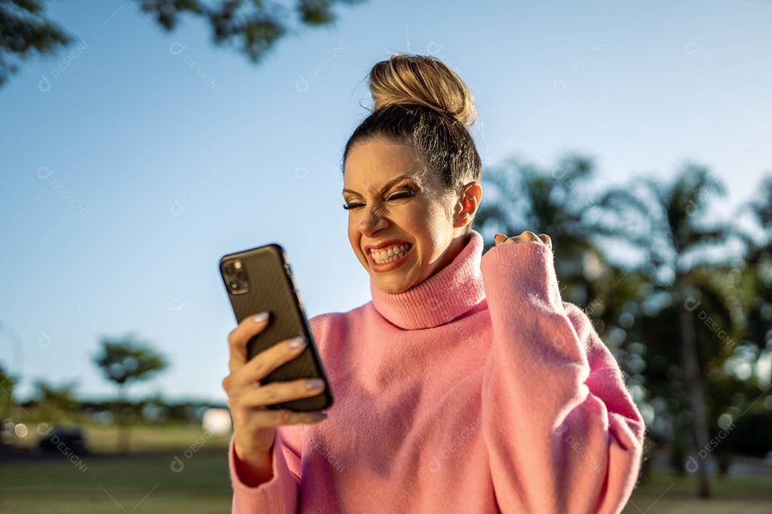 Imagem de uma jovem loira latina animada fazendo gesto de vencedor usando o celular no fundo do parque