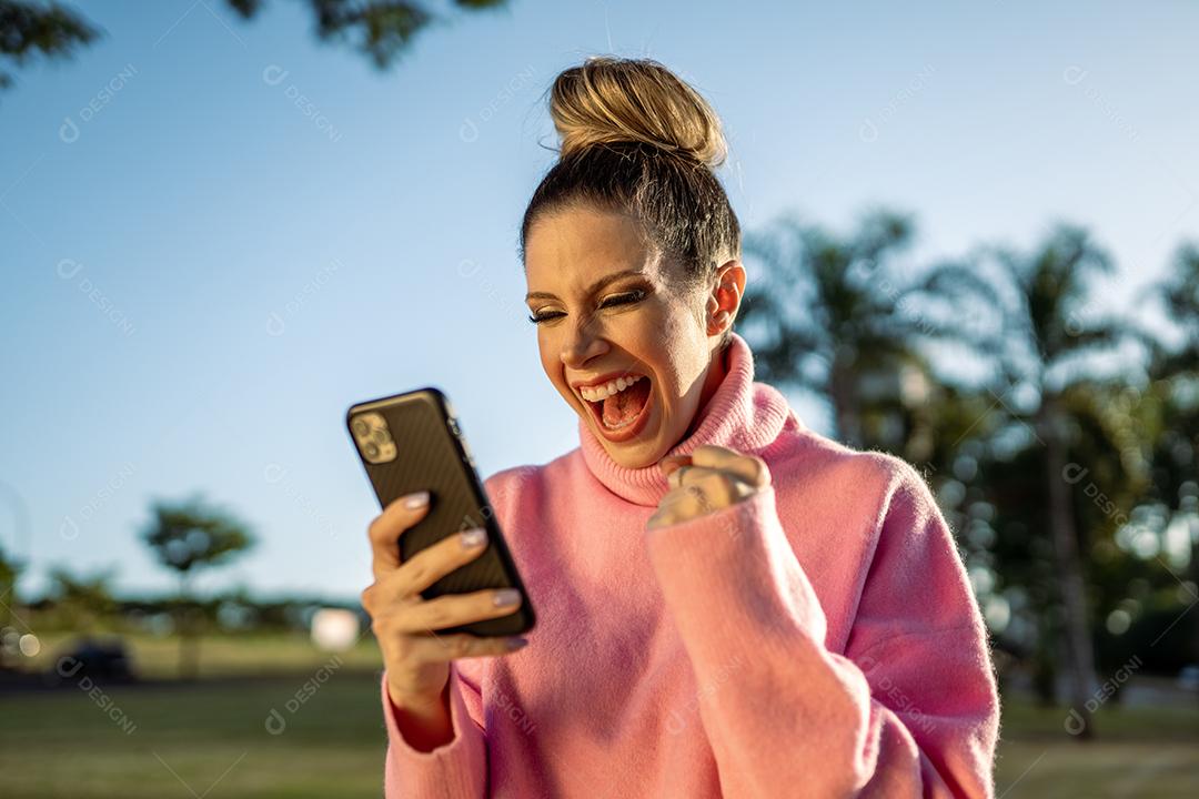 Imagem de uma jovem loira latina animada fazendo gesto de vencedor usando o celular no fundo do parque