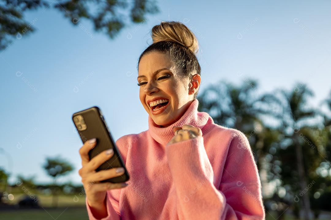 Imagem de uma jovem loira latina animada fazendo gesto de vencedor usando o celular no fundo do parque