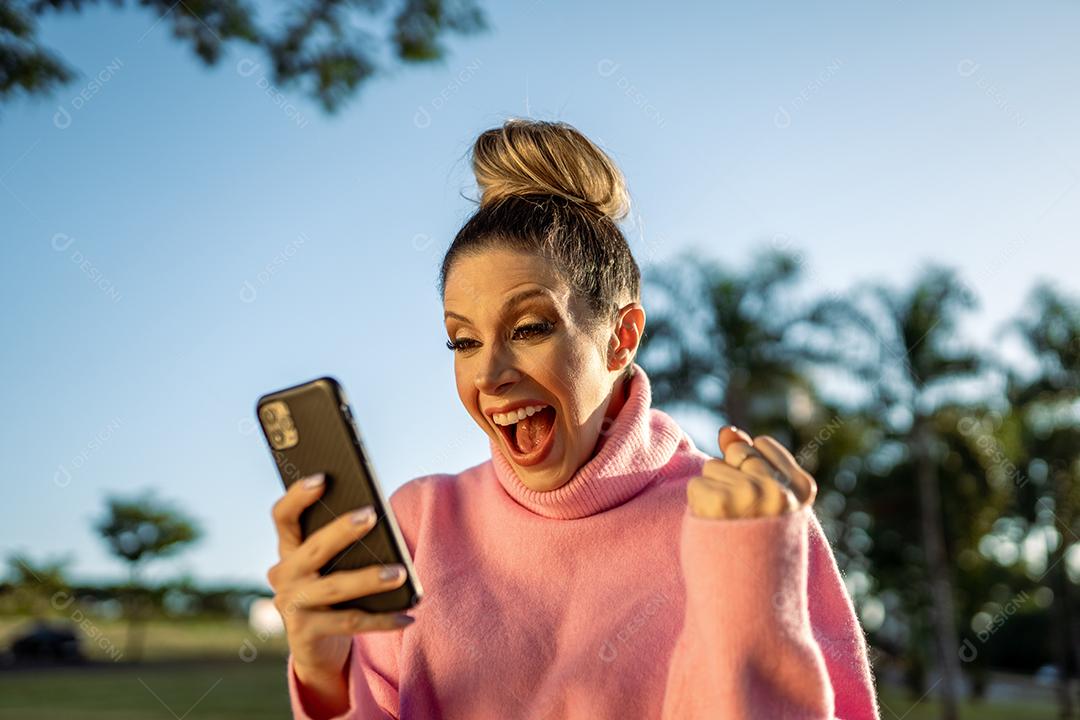 Imagem de uma jovem loira latina animada fazendo gesto de vencedor usando o celular no fundo do parque