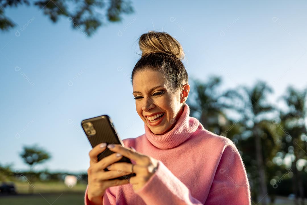 Imagem de uma jovem loira latina animada feliz usando o celular no fundo do parque