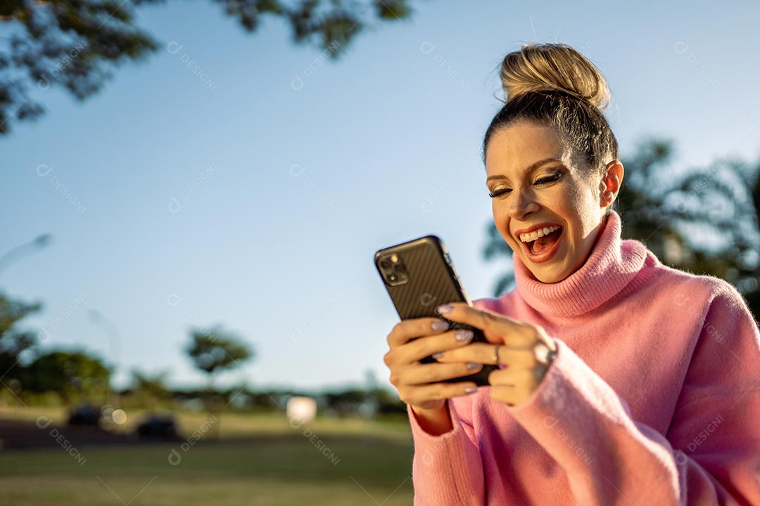 Imagem de uma jovem loira latina animada feliz usando o celular no fundo do parque