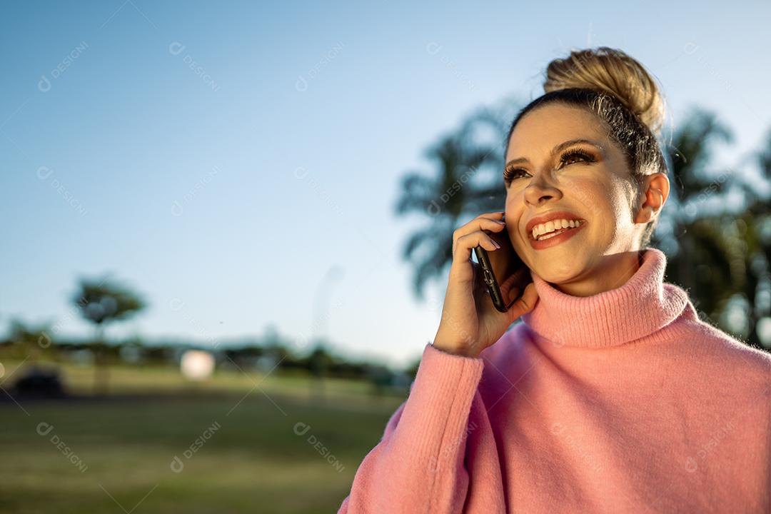 Imagem de uma jovem loira latina animada feliz usando o celular no fundo do parque