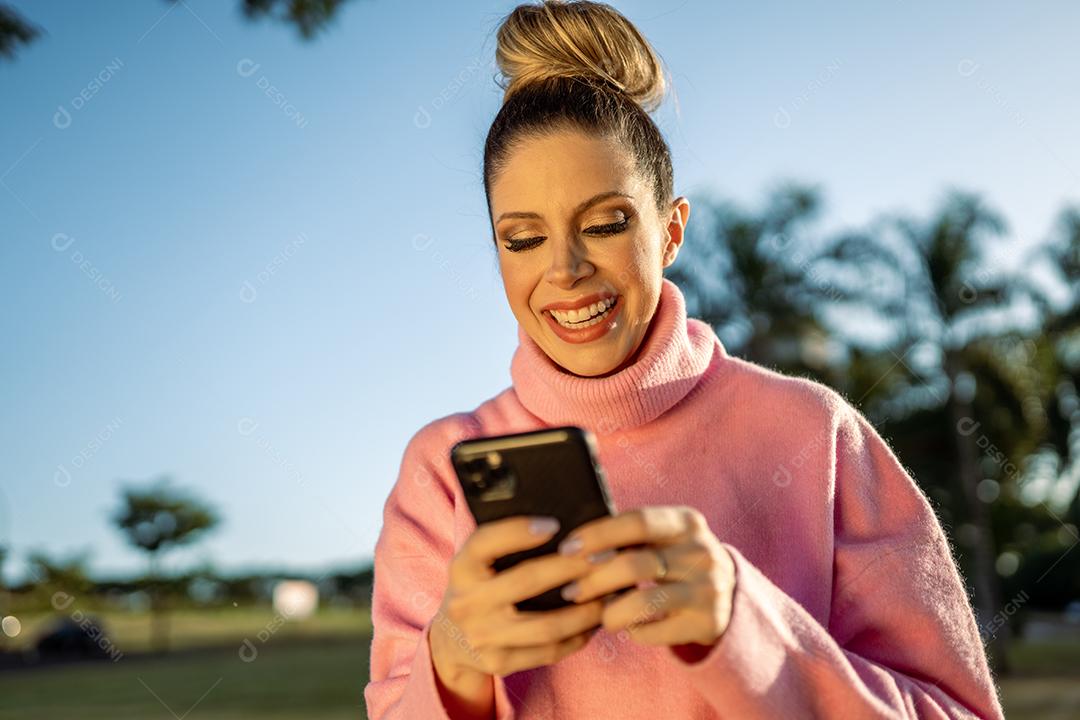 Imagem de uma jovem loira latina animada feliz usando o celular no fundo do parque