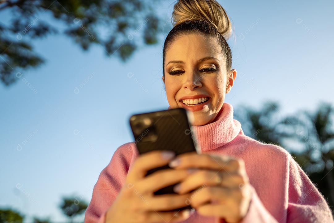 Imagem de uma jovem loira latina animada feliz usando o celular no fundo do parque