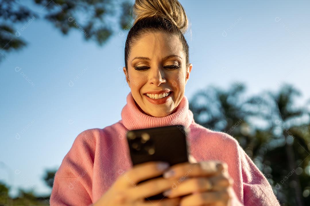 Imagem de uma jovem loira latina animada feliz usando o celular no fundo do parque