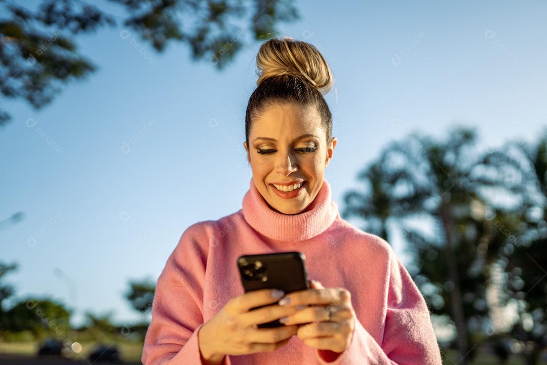 Imagem de uma jovem loira latina animada feliz usando o celular no fundo do parque