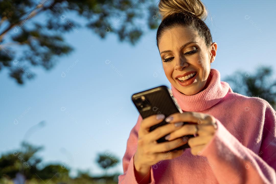 Imagem de uma jovem loira latina animada feliz usando o celular no fundo do parque