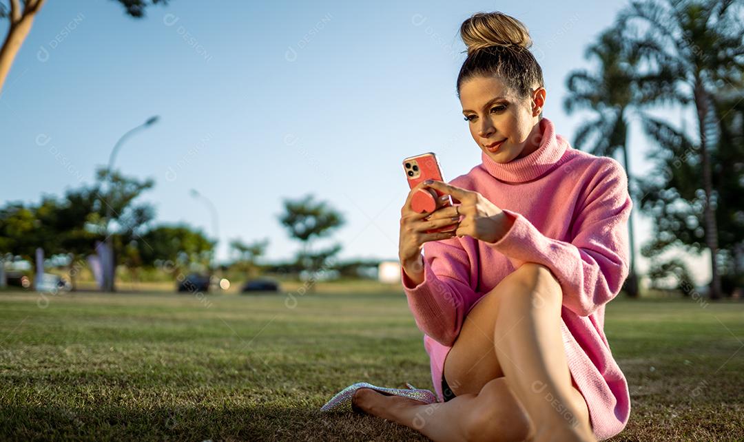 Imagem de uma jovem loira latina animada feliz usando o celular no fundo do parque