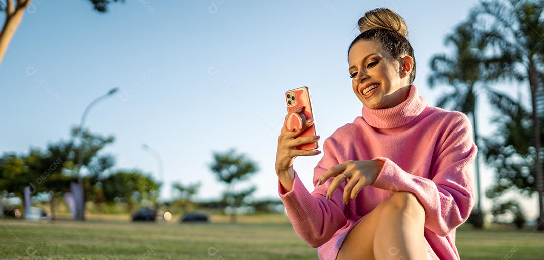 Imagem de uma jovem loira latina animada feliz usando o celular no fundo do parque