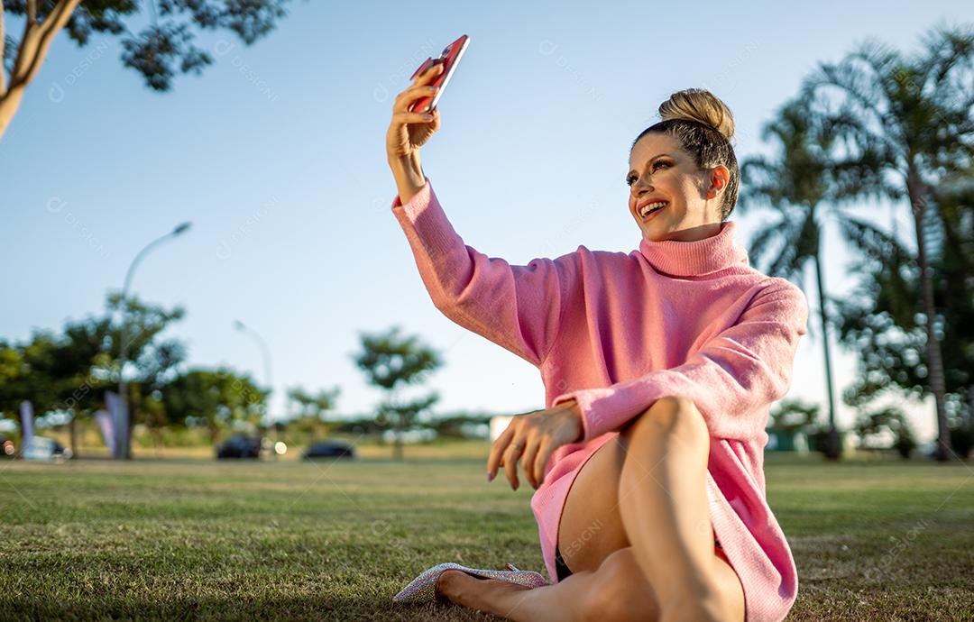 Imagem de uma jovem loira latina animada feliz fazendo selfie com celular no fundo do parque