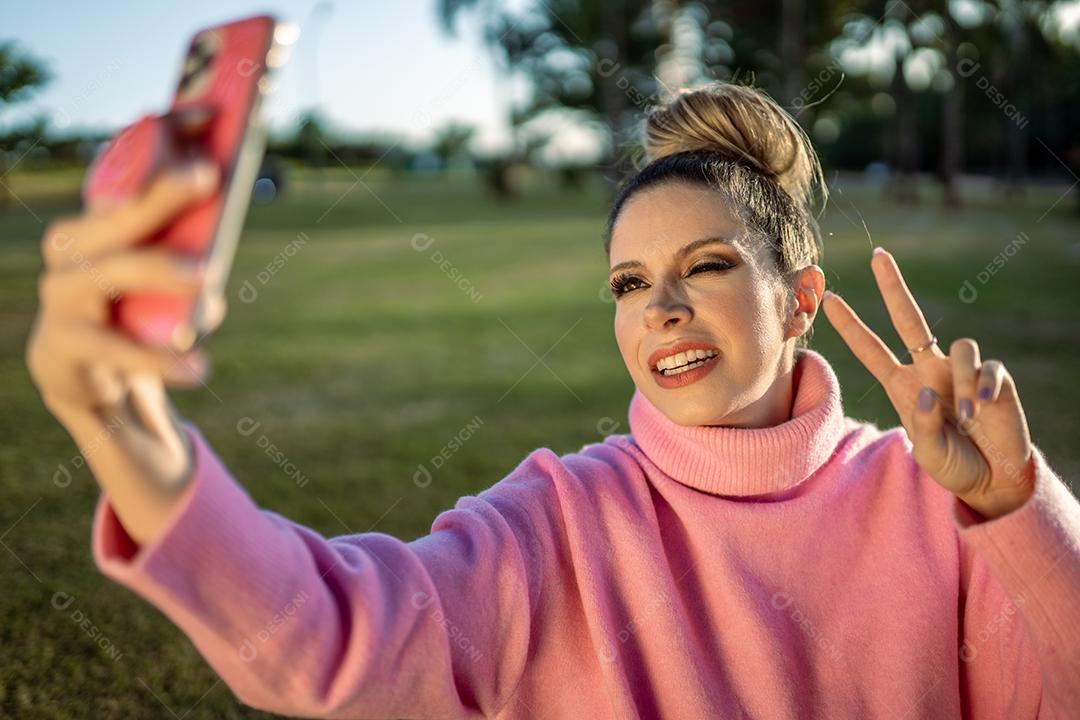 Imagem de uma jovem loira latina animada feliz fazendo selfie com celular no fundo do parque