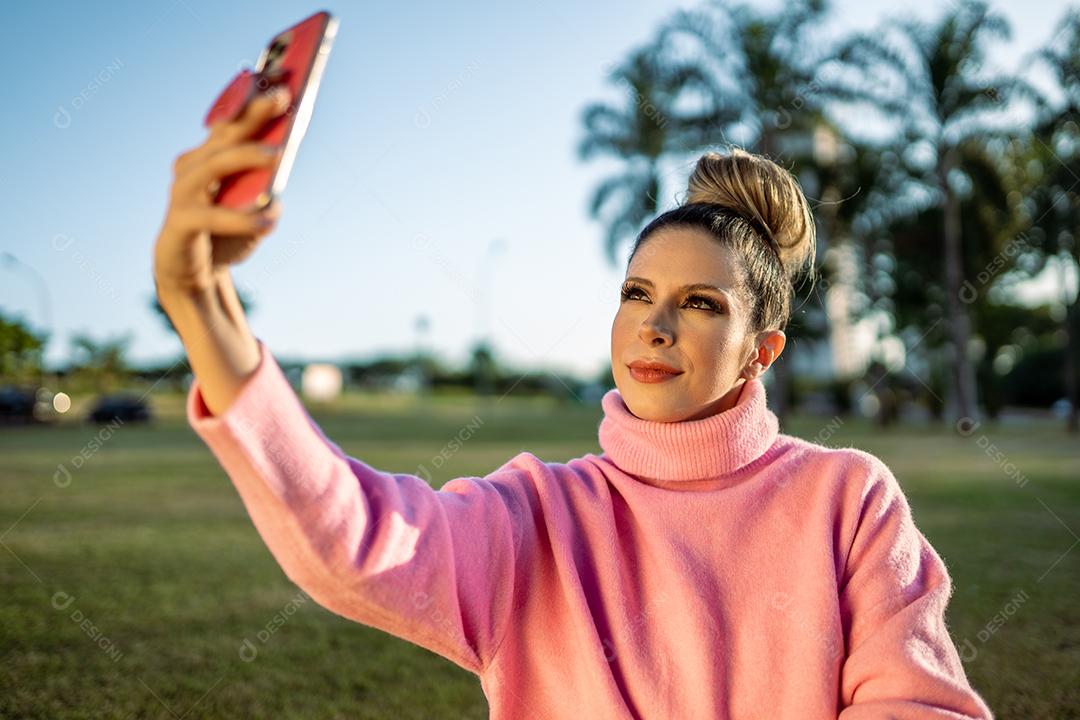 Imagem de uma jovem loira latina animada feliz fazendo selfie com celular no fundo do parque