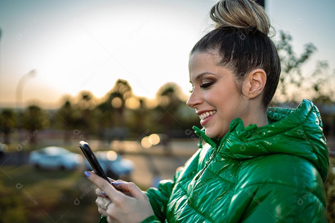 Imagem de uma jovem loira latina animada feliz usando o celular no fundo do parque