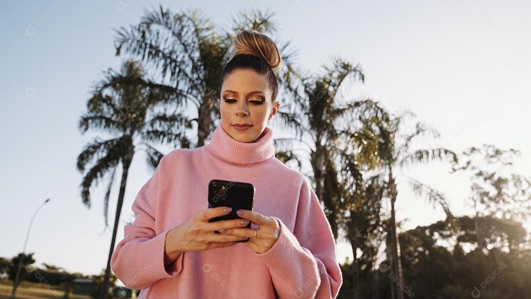 Imagem de uma jovem loira latina animada feliz usando o celular no fundo do parque