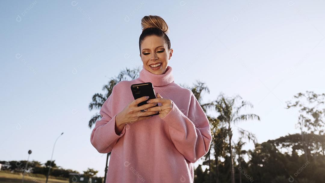 Imagem de uma jovem loira latina animada feliz usando o celular no fundo do parque