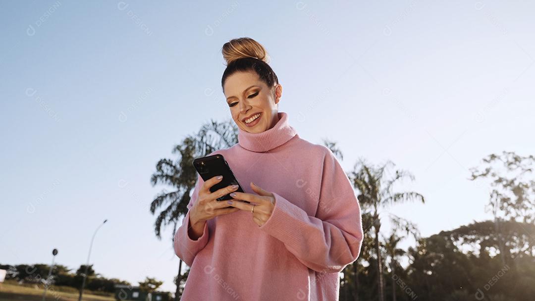 Imagem de uma jovem loira latina animada feliz usando o celular no fundo do parque