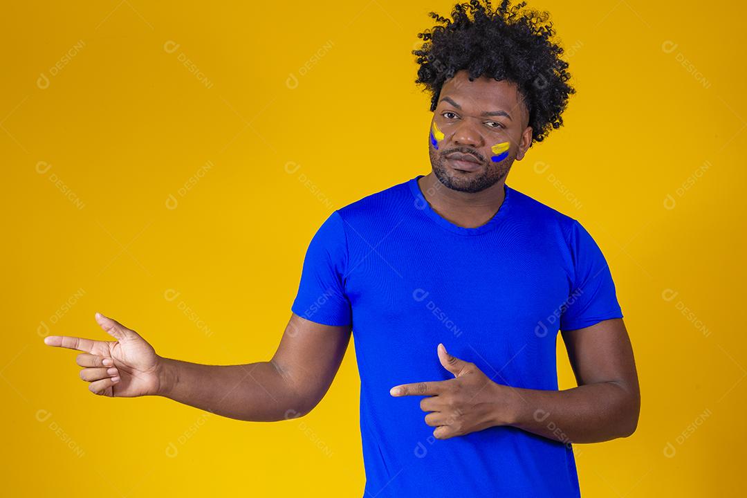 homem negro sobre fundo amarelo com rosto pintado com cores azuis e amarelas, apontando para o lado