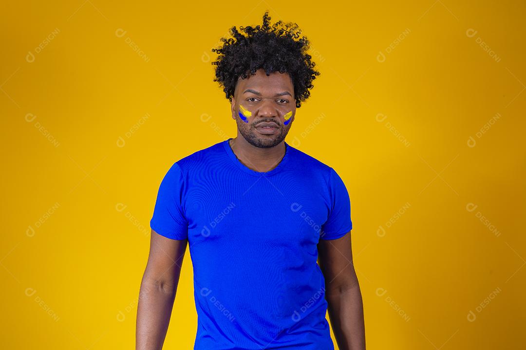 homem negro sobre fundo amarelo com o rosto pintado com cores azuis e amarelas se exibindo para a câmera