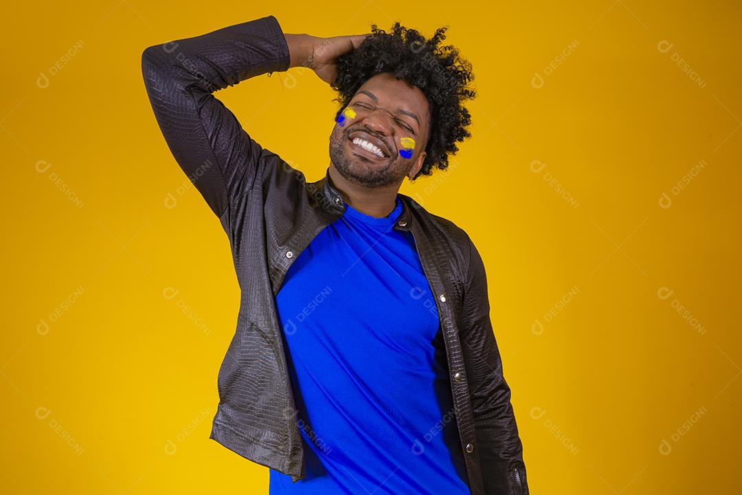 homem negro sobre fundo amarelo com o rosto pintado com cores azuis e amarelas se exibindo para a câmera